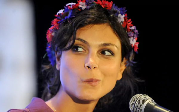 Morena Baccarin, a Brazilian actress, smiles charmingly while speaking at an event, adorned with a colorful flower crown. A vibrant HD background capturing her engaging presence.