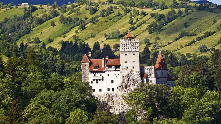 HD desktop wallpaper showcasing Bran Castle, a historic man-made fortress nestled among lush green hills and dense forest.