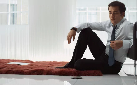 HD PC desktop wallpaper featuring English actor Clive Owen seated on the floor in a modern, light-filled room wearing a shirt and tie.