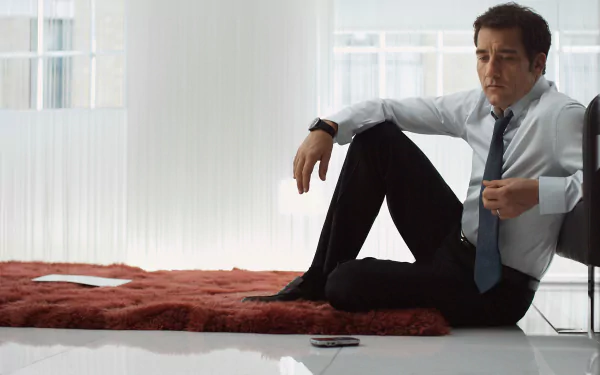 HD PC desktop wallpaper featuring English actor Clive Owen seated on the floor in a modern, light-filled room wearing a shirt and tie.