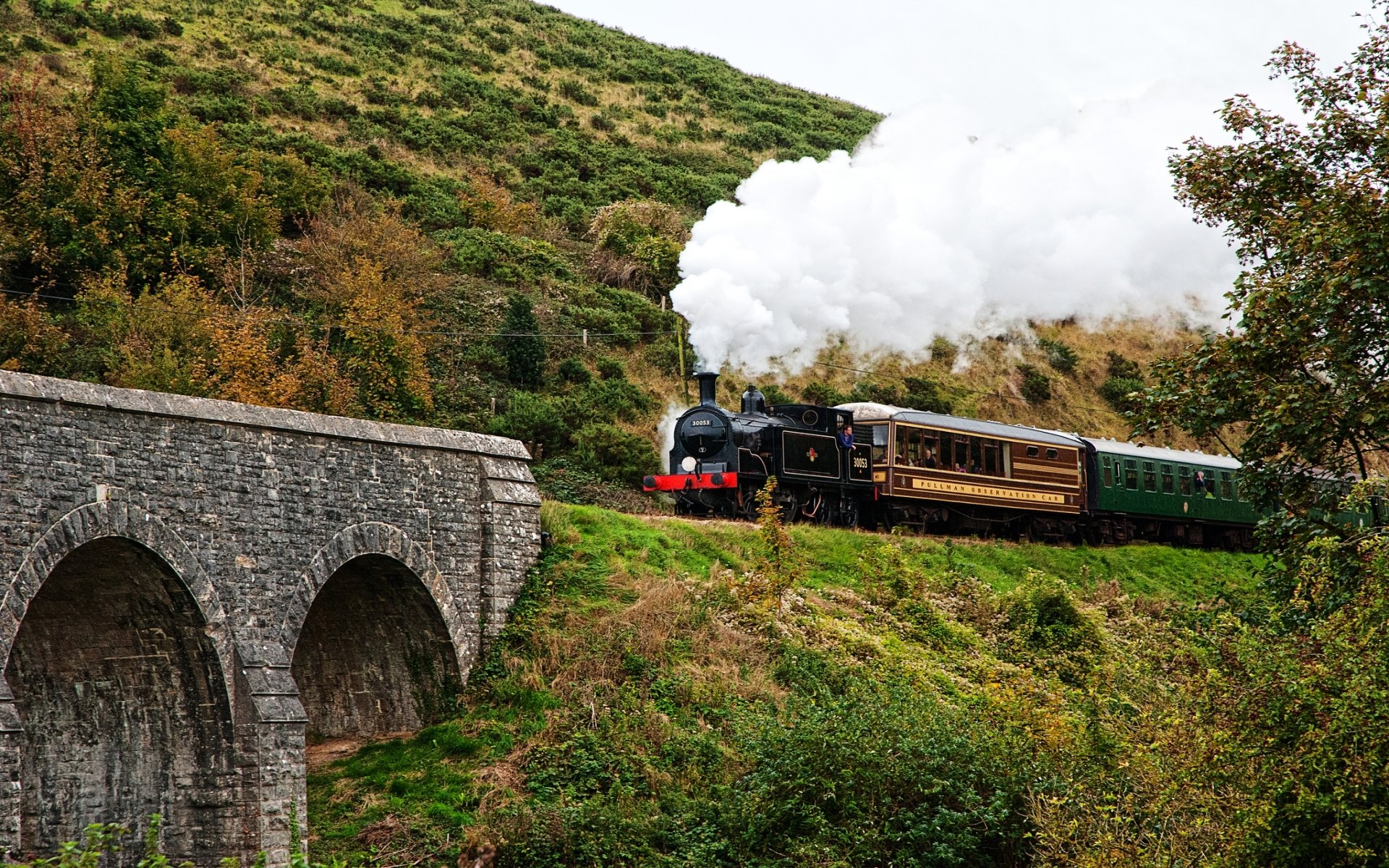 HD desktop wallpaper featuring a classic steam train crossing a stone bridge surrounded by lush green hills and trees under a cloudy sky.