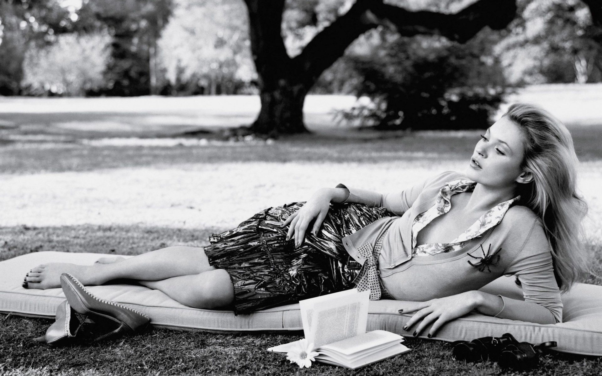 Black-and-white 2K Quad HD PC desktop wallpaper of a reclining blonde fashion model in a casual outfit on grass beneath a tree, sandals and open books beside her in a serene park scene.