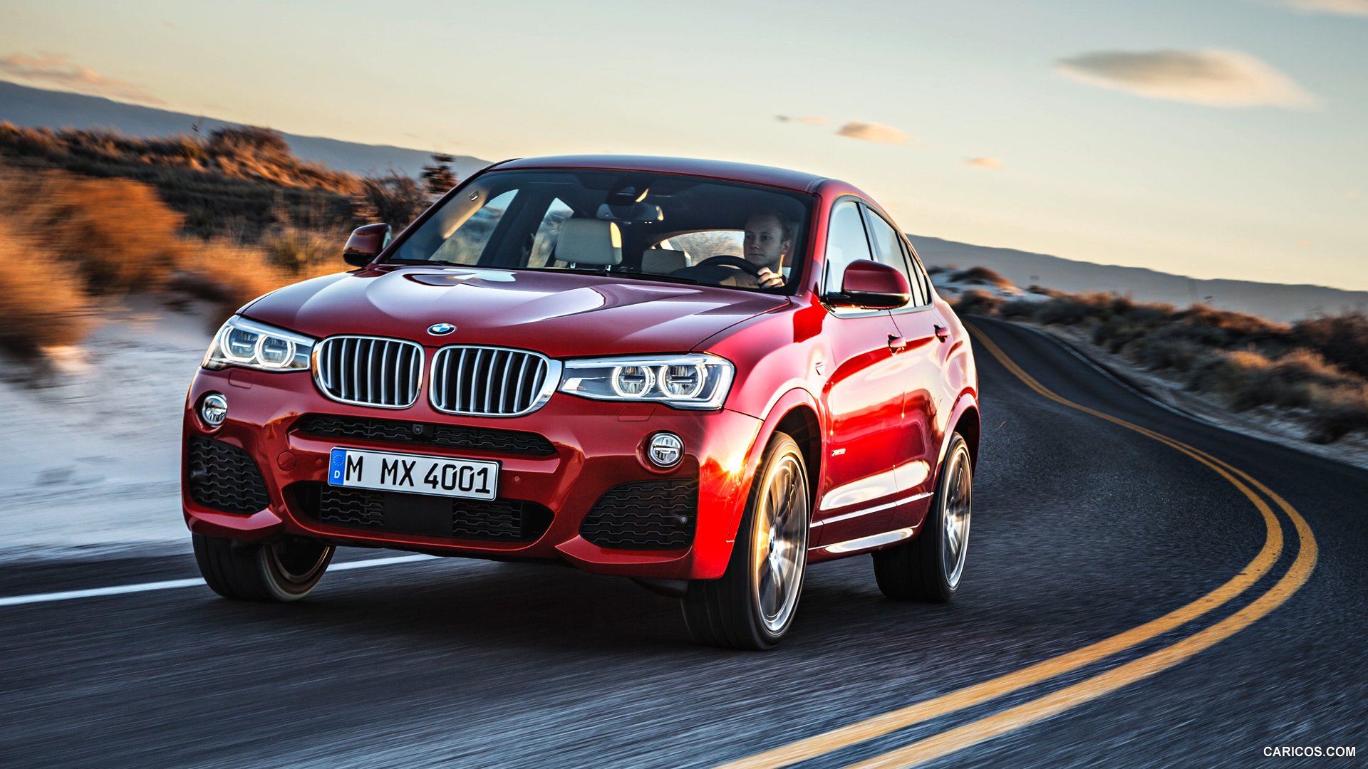 HD PC desktop wallpaper featuring a red BMW X4 driving on a winding road through a desert landscape at sunset.