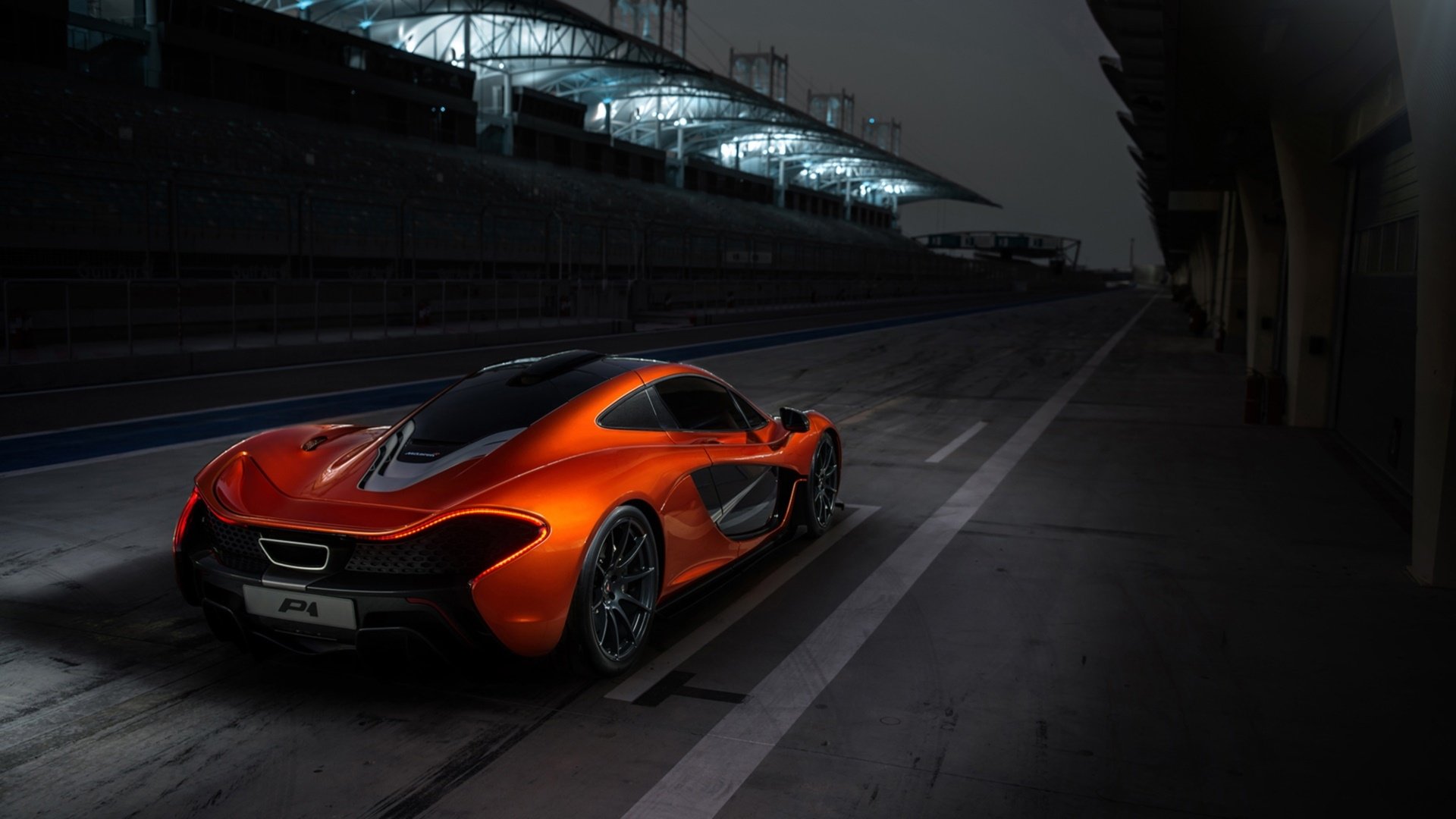 HD PC desktop wallpaper of a glossy orange McLaren P1 sports car parked in a dark racetrack pit lane, dramatic overhead lights reflecting on its aerodynamic body.