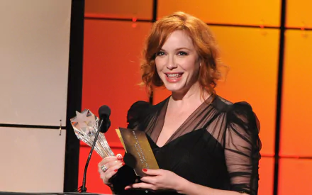 HD desktop wallpaper of American actress and celebrity Christina Hendricks smiling on stage, holding a bouquet and an envelope against an orange backdrop.