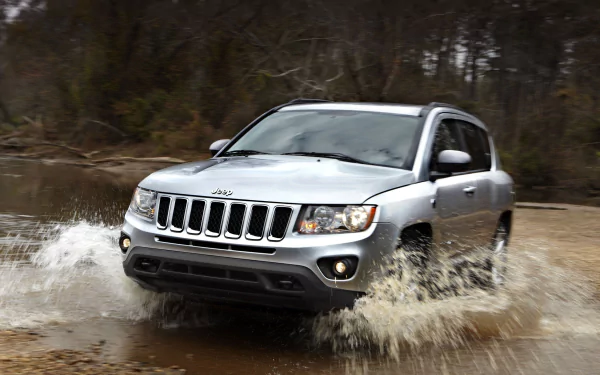 Silver Jeep Compass splashing through shallow water, dynamic 2K Quad HD PC desktop wallpaper background of an off-road vehicle in a wooded setting.