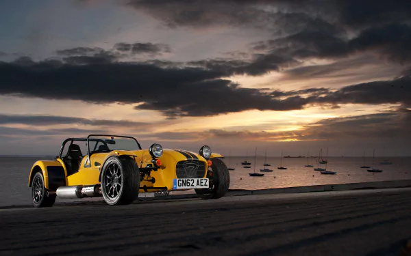 Yellow Caterham Supersport R vehicle parked on a beach at sunset with moored boats on calm water — 2K Quad HD PC desktop wallpaper background.