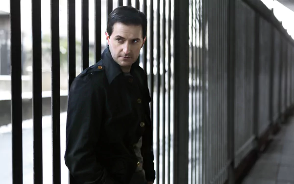 2K Quad HD PC desktop wallpaper of an English actor in a dark coat leaning by metal railings, moody cinematic portrait with cool, desaturated tones.