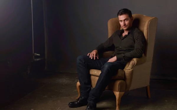 HD desktop wallpaper featuring English actor and celebrity Richard Armitage seated confidently in an armchair against a dark background.