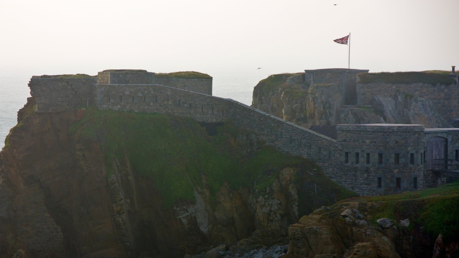 A stunning HD wallpaper featuring Fort Clonque, showcasing its impressive stone architecture perched on cliffs with a flag waving against a misty backdrop.