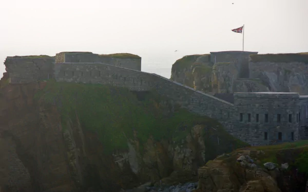 A stunning HD wallpaper featuring Fort Clonque, showcasing its impressive stone architecture perched on cliffs with a flag waving against a misty backdrop.