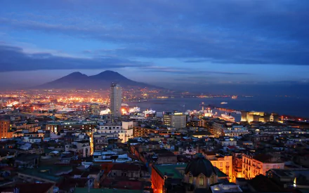 Nighttime cityscape of Naples, Italy, featuring illuminated buildings and the silhouette of Mount Vesuvius in the background under a twilight sky.