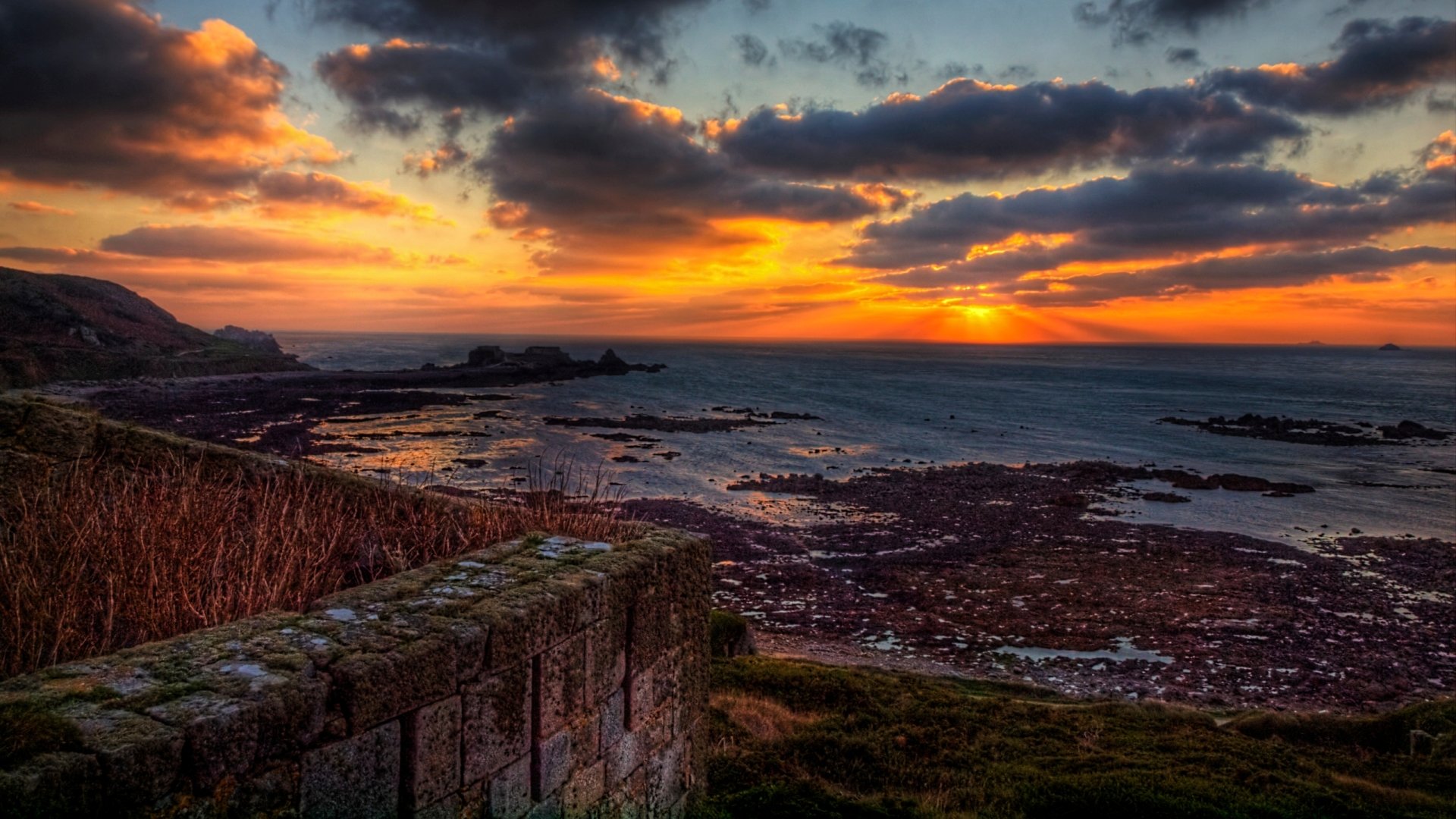 Sunset over rocky coast with man-made Fort Tourgis stone ramparts in foreground, dramatic clouds and tidal pools — 2K Quad HD PC desktop wallpaper/background.