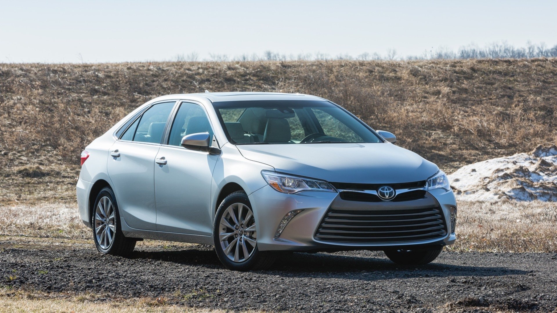 HD PC desktop wallpaper of a silver 2015 Toyota Camry vehicle parked on gravel by a grassy embankment under a clear sky.