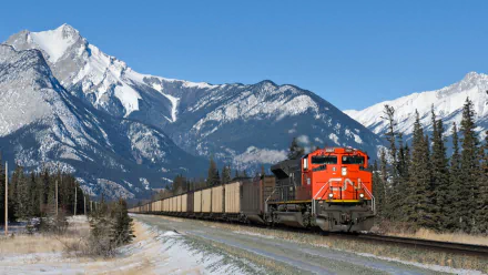 HD desktop wallpaper featuring a bright orange freight train traveling through a snowy mountainous landscape under a clear blue sky.