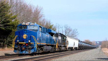 A blue diesel locomotive pulls a long train of black freight cars along a rural track, surrounded by trees and a clear sky. An engaging HD desktop wallpaper for train enthusiasts.