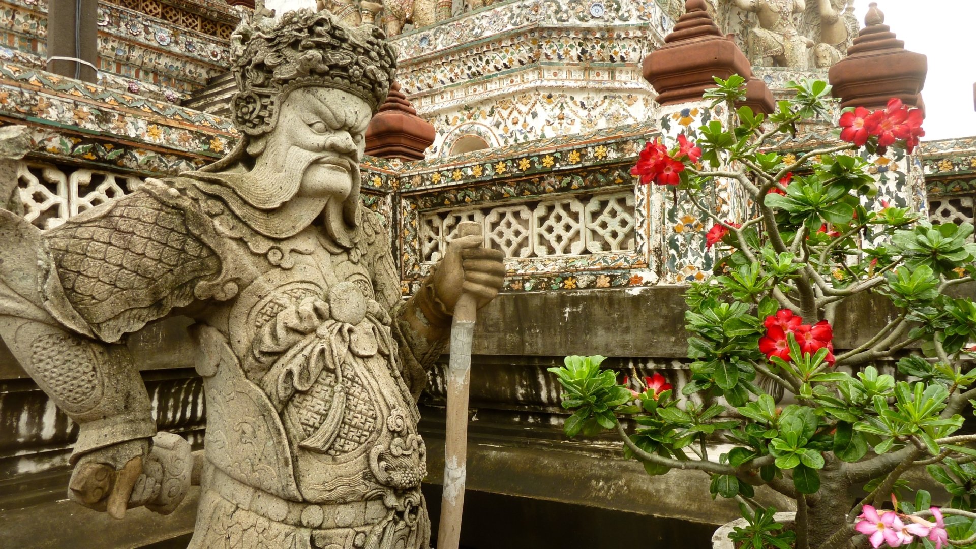 Religious view of Wat Arun Temple: carved stone guardian beside red blooms and ornate spires — 2K Quad HD PC desktop wallpaper and background.