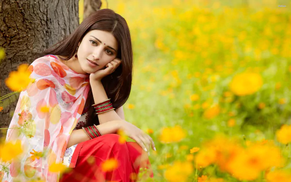 A serene Shruti Haasan poses under a tree, surrounded by vibrant yellow flowers, wearing traditional attire. This HD image serves as a stunning desktop wallpaper.