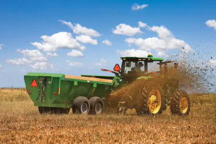 HD PC desktop wallpaper showing a John Deere vehicle spreading organic material across a field under a blue sky with scattered clouds.