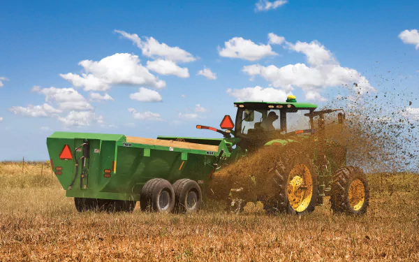 HD PC desktop wallpaper showing a John Deere vehicle spreading organic material across a field under a blue sky with scattered clouds.