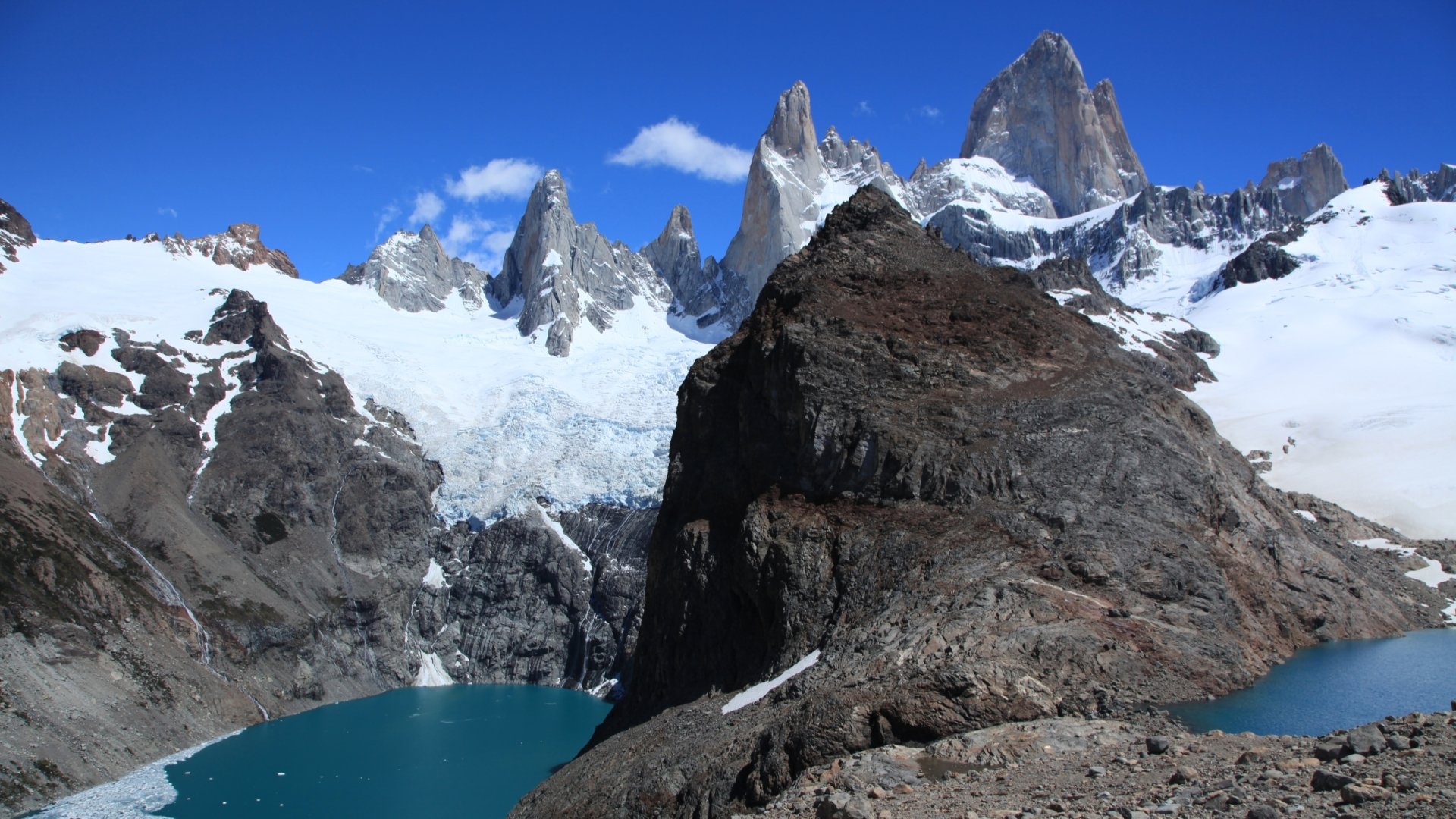 HD desktop wallpaper showcasing the striking snowy peaks of Mount Fitz Roy against a clear blue sky, surrounded by rugged rocky terrain and a turquoise glacial lake.