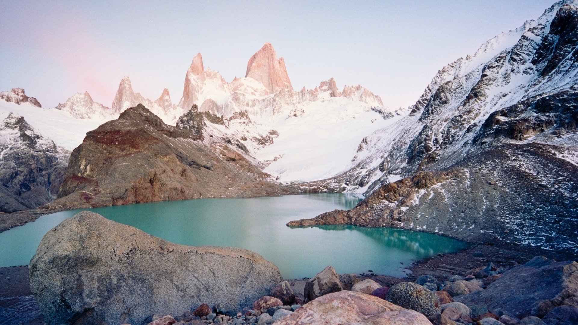 Nature HD PC desktop wallpaper and background: Mount Fitz Roy rising above a turquoise glacial lake, rocky foreground and snow-capped peaks beneath a pastel sunrise sky.