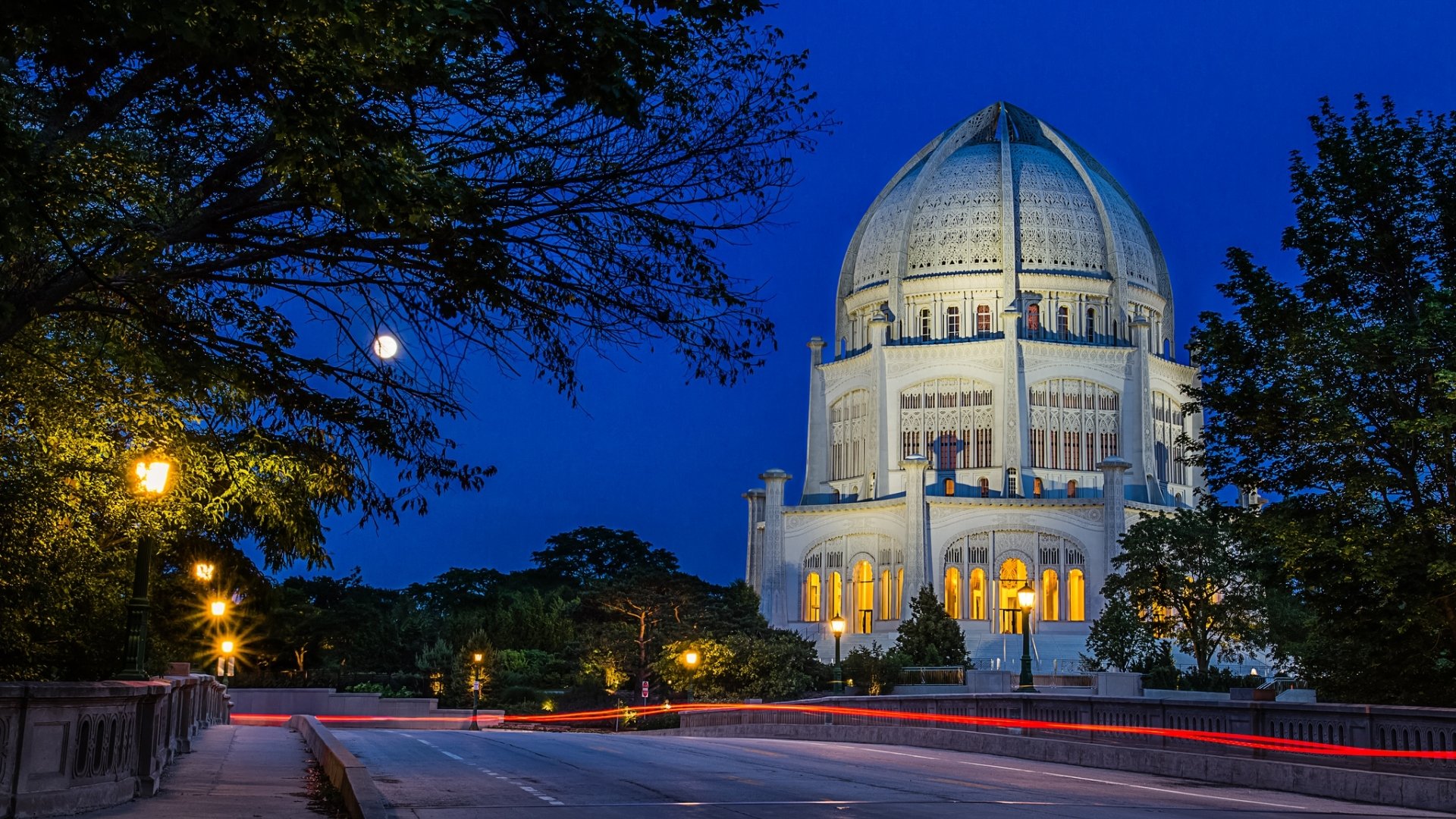 HD PC desktop wallpaper background of an illuminated Baha'i Temple at dusk, moonlit deep-blue sky, surrounding trees and streaking light trails along a quiet road.
