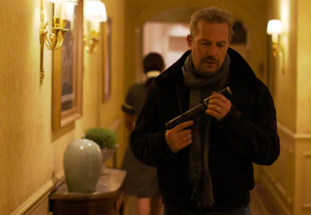 HD PC desktop wallpaper from the movie 3 Days to Kill: a man in a coat and scarf inspects a handgun while walking down a hotel hallway.