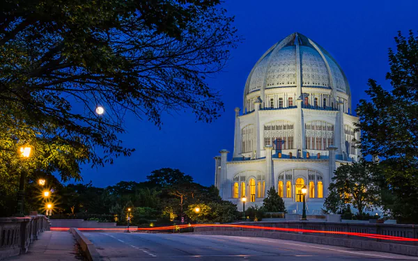 HD PC desktop wallpaper background of an illuminated Baha'i Temple at dusk, moonlit deep-blue sky, surrounding trees and streaking light trails along a quiet road.