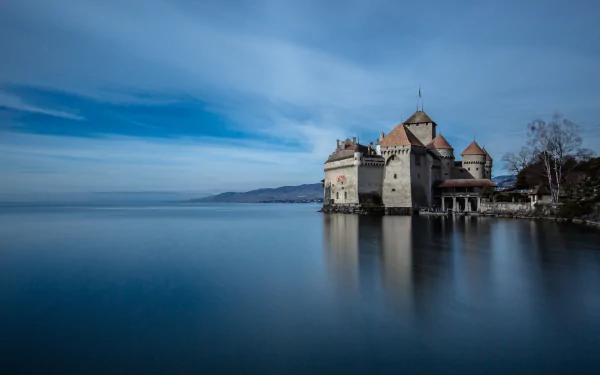 A serene HD desktop wallpaper featuring Chateau de Chillon, a historic man-made castle in Veytaux, Switzerland, reflected on calm waters under a blue sky.