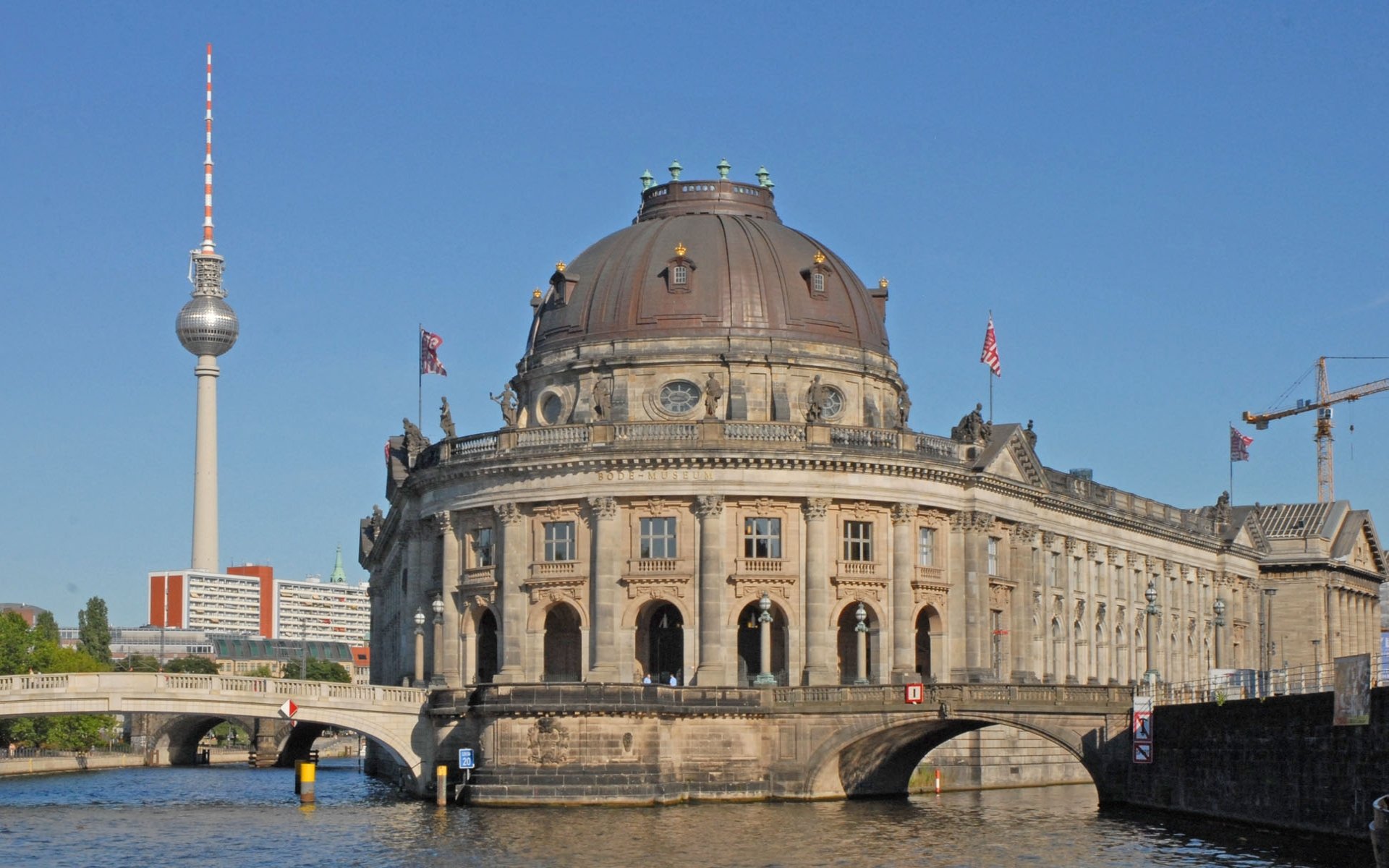 HD desktop wallpaper showcasing the Bode Museum, a grand man-made historic building by the river, with Berlin's TV tower visible in the clear blue sky.