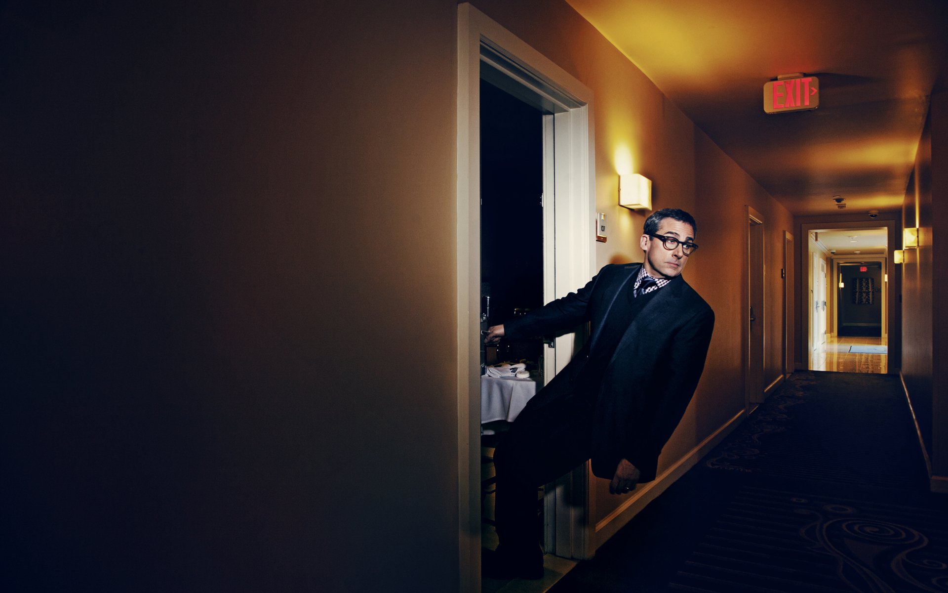 Man in suit and glasses peeks from a hotel room into a dimly lit corridor; cinematic HD PC desktop wallpaper.