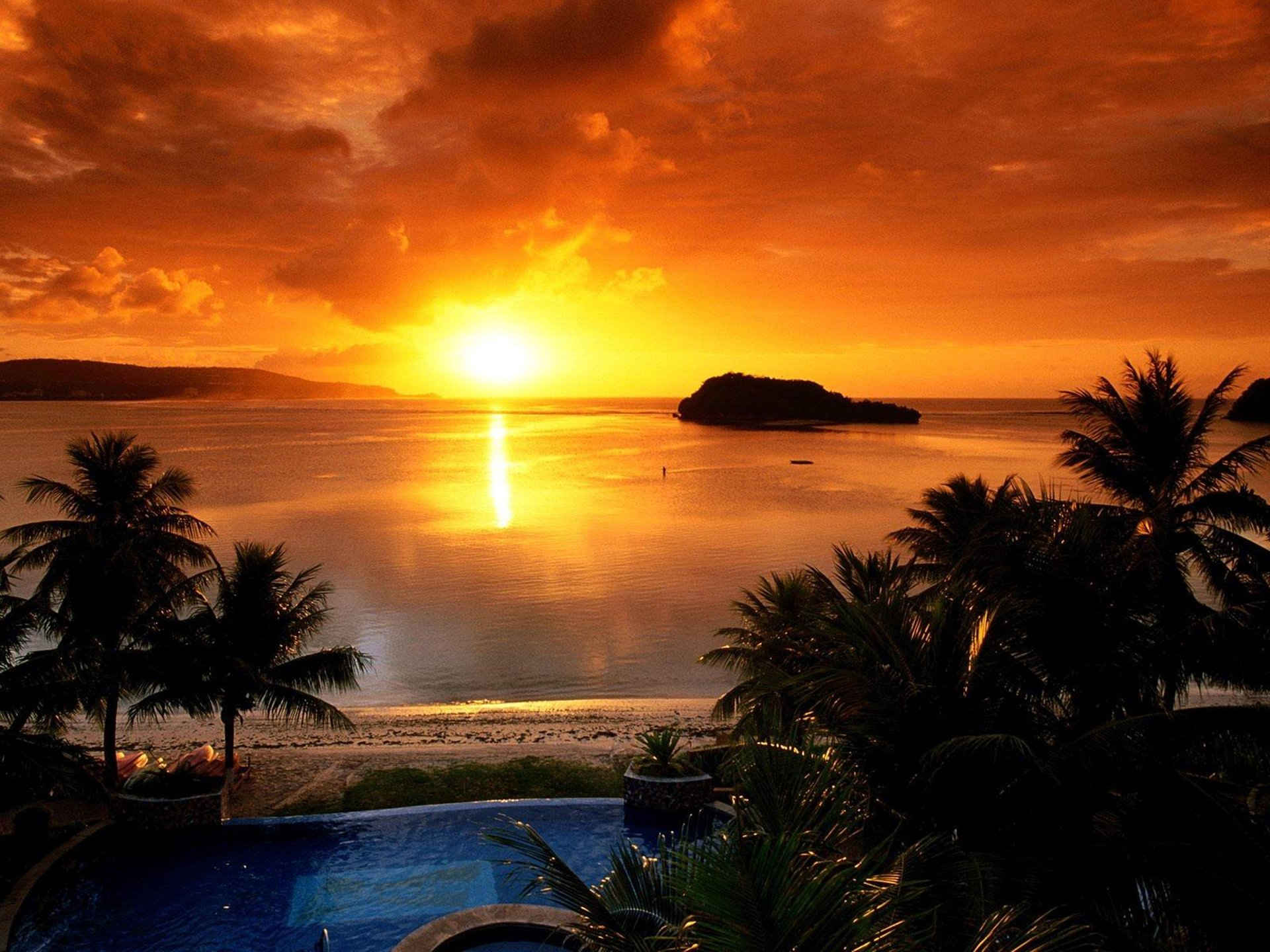 A stunning sunset illuminates the sky with vibrant orange hues, reflecting off calm waters beside a sandy beach and palm trees, creating a serene nature scene for a HD desktop wallpaper.