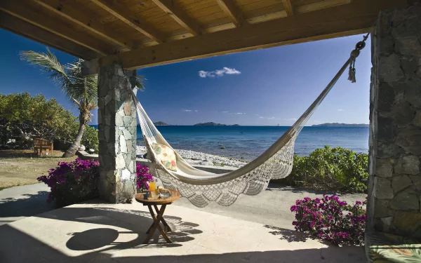 HD PC desktop wallpaper showing a man-made beachfront patio with a woven hammock strung between stone pillars, a small table with flowers, and turquoise ocean and sandy beach beyond.