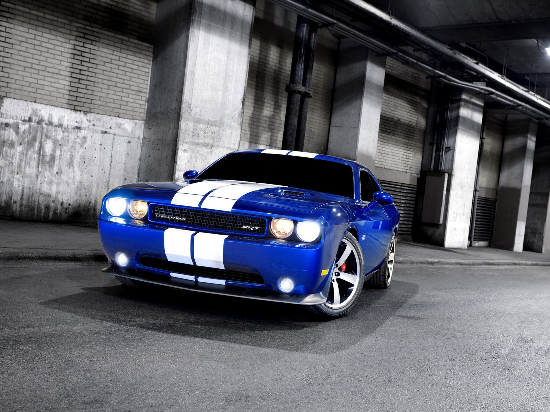 HD PC desktop wallpaper: blue Dodge Challenger SRT8 muscle vehicle with white racing stripes, headlights on, parked in a dim industrial garage.
