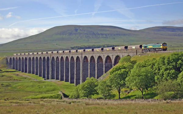HD PC desktop wallpaper of the man-made Ribblehead Viaduct crossing green moors with a passing freight train under a bright blue sky.