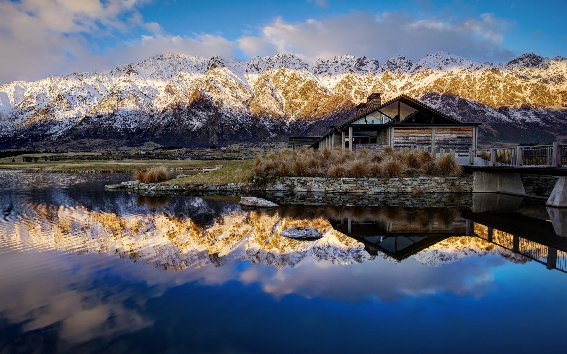 HD desktop wallpaper of Lake Wakatipu in Queenstown, New Zealand, showcasing serene waters reflecting snow-capped mountains under a partly cloudy sky.