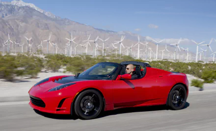 A red Tesla Roadster driving with wind turbines in the background, as a sleek HD desktop wallpaper.