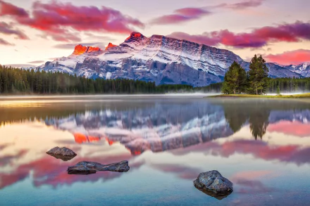 HD PC desktop wallpaper: serene nature scene of Two Jack Lake at sunrise, snow-capped mountains and vivid clouds mirrored in the calm water with foreground rocks and a pine treeline.