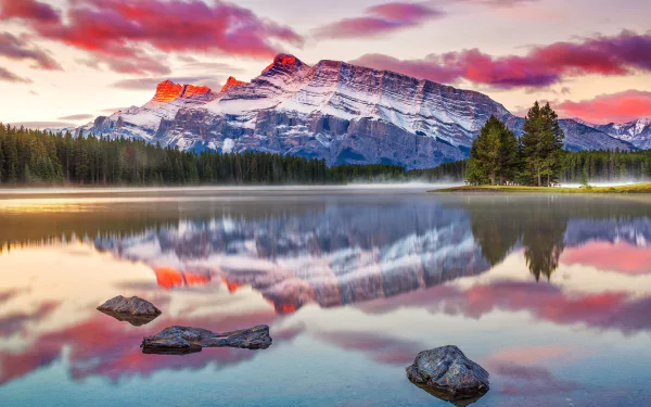 HD PC desktop wallpaper: serene nature scene of Two Jack Lake at sunrise, snow-capped mountains and vivid clouds mirrored in the calm water with foreground rocks and a pine treeline.