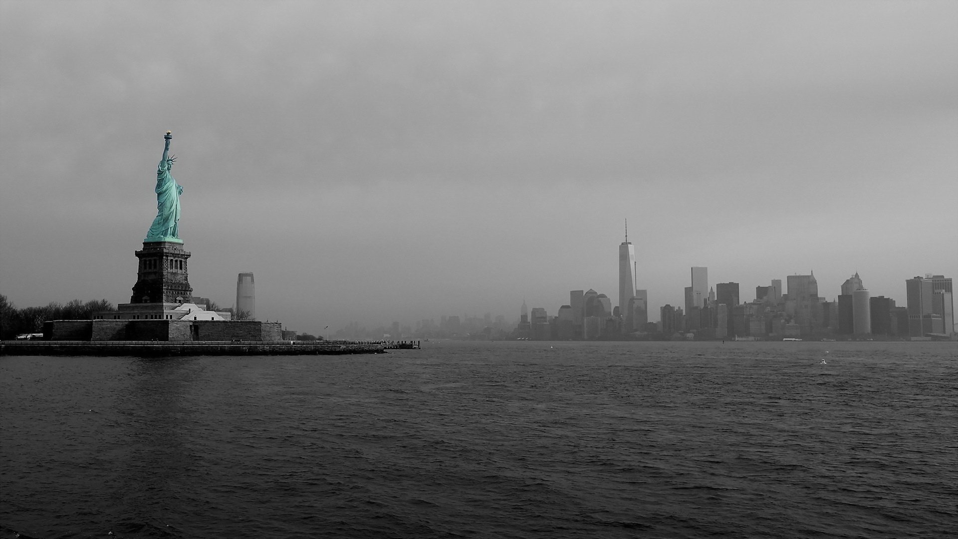 HD PC desktop wallpaper: black-and-white view of the man-made Statue of Liberty on Liberty Island, overlooking water toward a foggy Manhattan skyline.