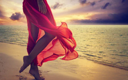 A woman in a flowing red dress stands barefoot on a sandy beach at sunset, with the ocean stretching out under a colorful sky.