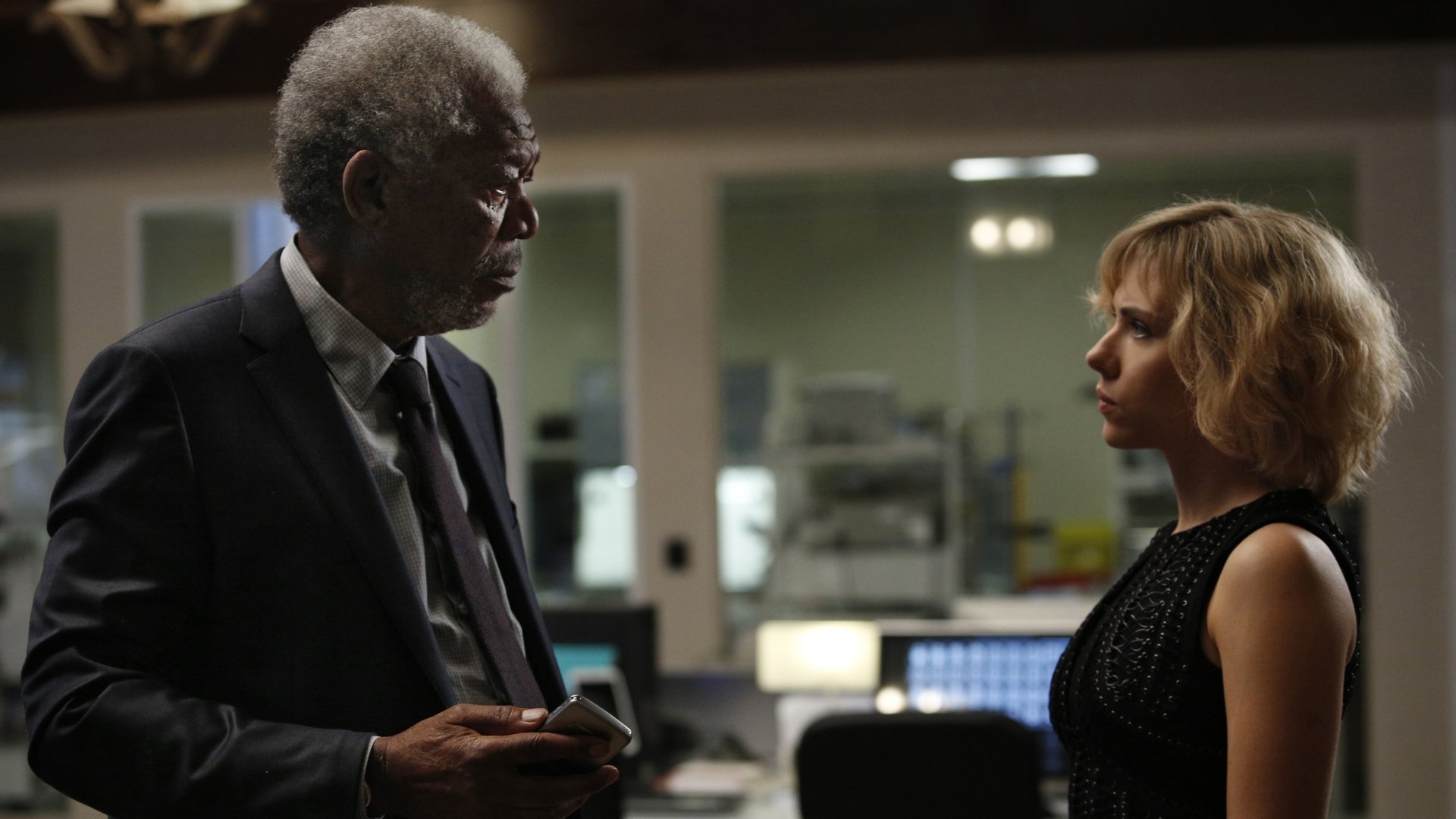 HD desktop wallpaper featuring Morgan Freeman and Scarlett Johansson in a scene from the movie Lucy, showcasing intense conversation in a dimly lit room.