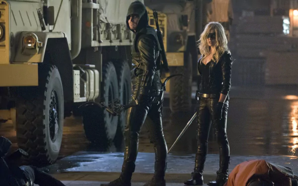Caity Lotz as Black Canary and Stephen Amell as Arrow stand in a dramatic scene, showcasing their characters' intensity from the TV show Arrow. The backdrop features military vehicles.
