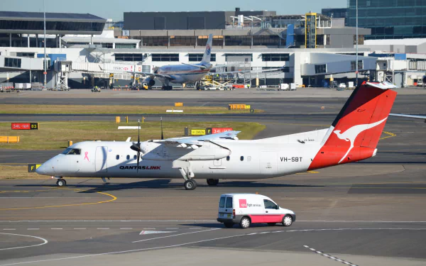 Bombardier Q315 aircraft in QantasLink livery taxiing at Sydney airport, with terminal buildings and a ground service vehicle in the background.