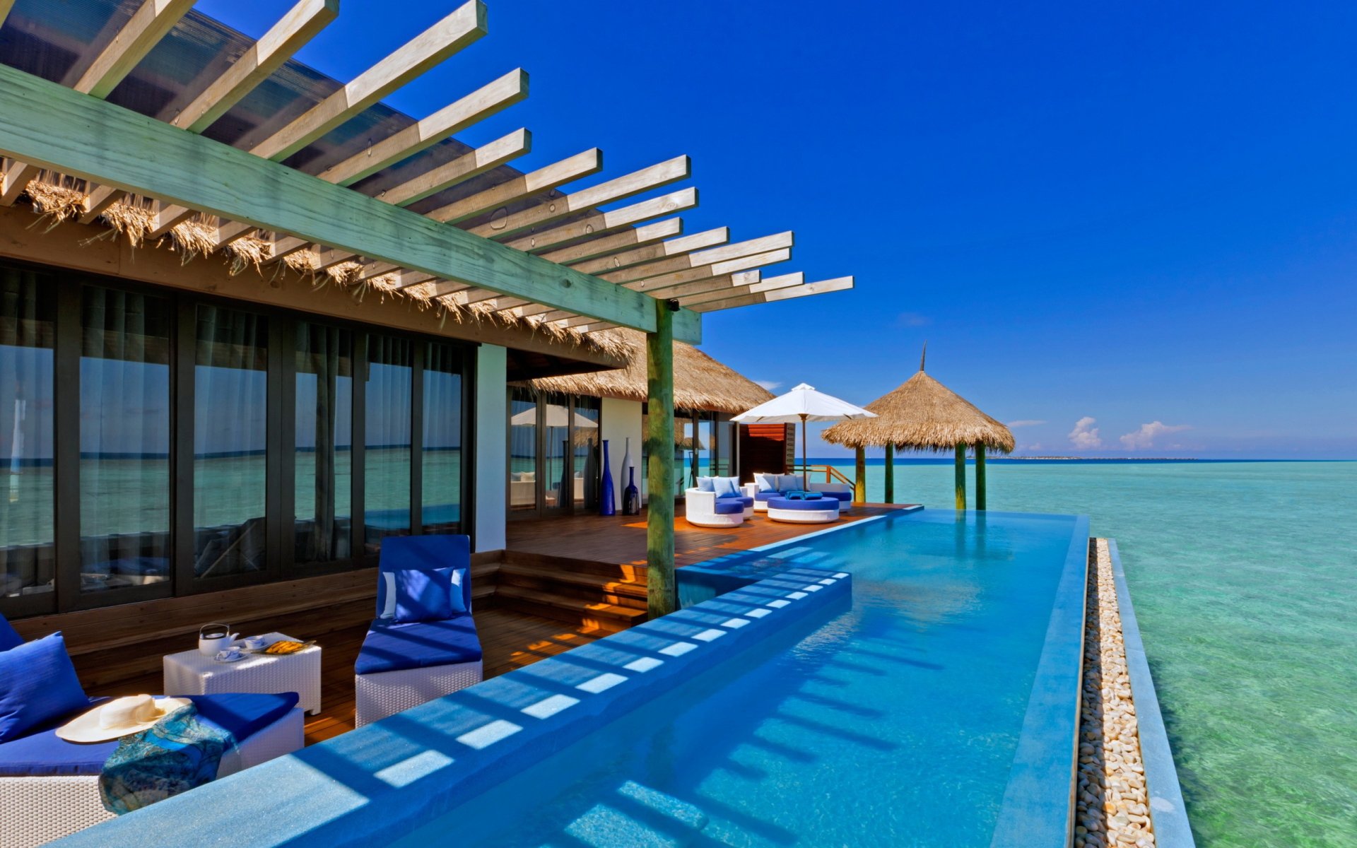2K Quad HD PC desktop wallpaper showing a man-made overwater resort with an infinity pool, lounge seating and thatched cabanas over turquoise sea beneath a clear blue sky.