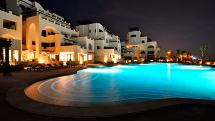 HD desktop wallpaper of a man-made pool at night, illuminated with blue lights, surrounded by modern white buildings and lounge chairs.