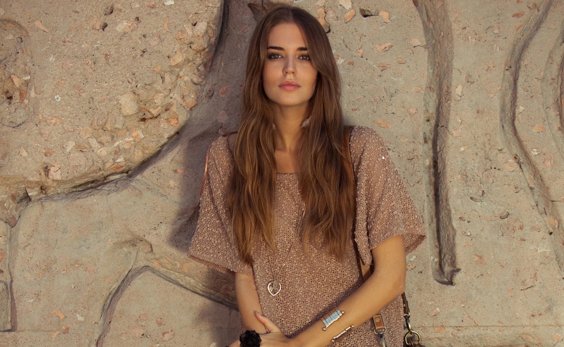 HD desktop wallpaper featuring Spanish woman Clara Alonso standing against a textured stone wall, wearing a brown knit top and long loose hair.