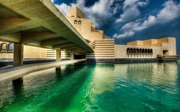 HD wallpaper of a museum in Doha, Qatar, featuring striking man-made architecture over a calm, green body of water with a dramatic sky in the background.