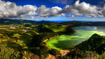A panoramic view of a vibrant lake surrounded by lush forests and rolling hills under a dramatic sky with clouds, showcasing the stunning landscape of the Azores, Portugal.